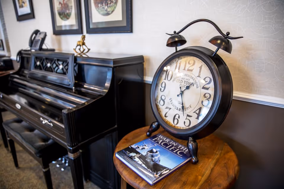 A black upright piano with a cushioned bench is positioned against a wall with framed artwork above it. Next to the piano, on a round wooden table, sits a large vintage-style black alarm clock and a book titled 'War in the Pacific.'