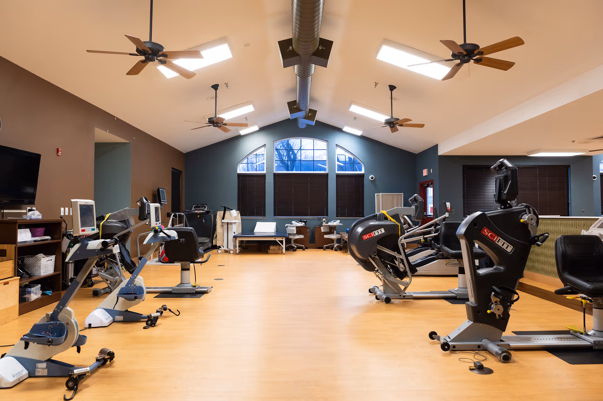 A spacious exercise room with several stationary exercise bikes and fitness equipment arranged on a light wood floor. The room has high ceilings with ceiling fans and skylights, large arched windows with blinds, and a TV mounted on the wall. There are also some chairs and tables along the walls.