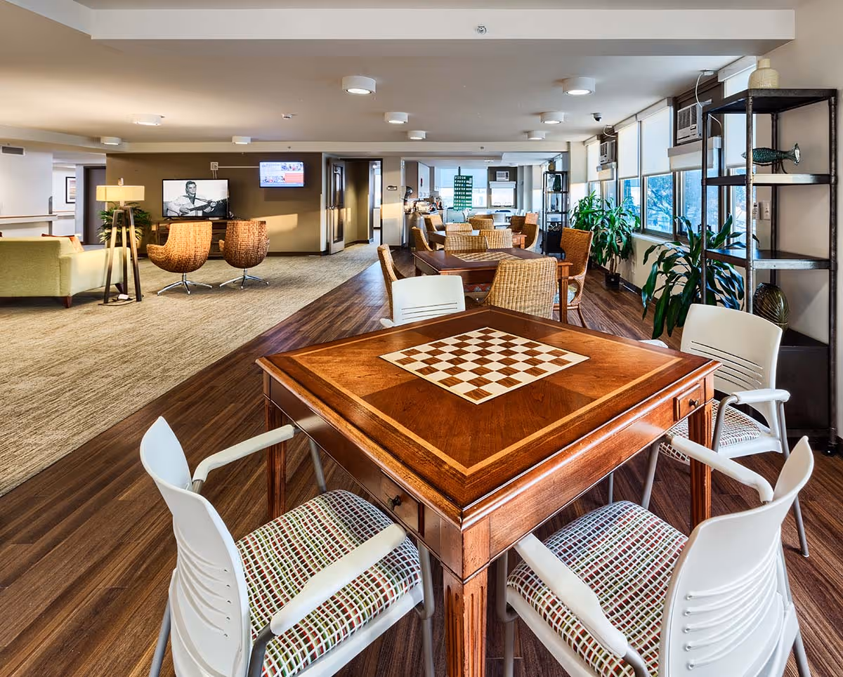 A spacious, well-lit common area in Kalākaua Gardens featuring a wooden game table with a built-in chessboard surrounded by four white chairs with patterned cushions. In the background, there are multiple wicker chairs and tables, large windows with natural light, potted plants, a television mounted on the wall, and a cozy seating area with a green sofa and two wicker chairs.