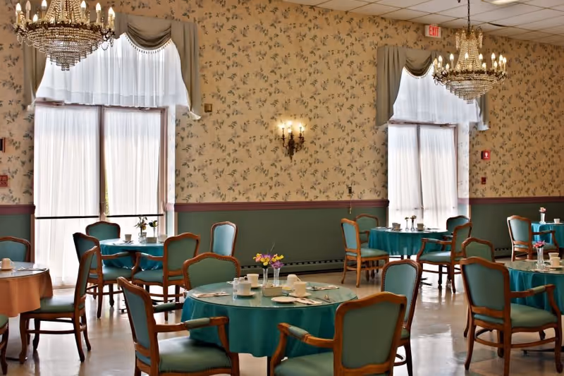 Elegant dining room with round tables draped in teal tablecloths, wooden chairs, chandeliers, and floral wallpapered walls.