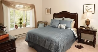 A cozy bedroom with a large bed featuring a dark wooden headboard and blue-gray bedding. There are multiple pillows on the bed, a nightstand with a lamp and decorative items, a window with sheer curtains letting in natural light, and a small chair with a throw blanket in the corner. The room has a warm and inviting atmosphere.