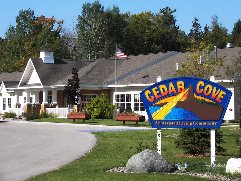 Exterior view of Cedar Cove Assisted Living community building with a large colorful sign in the foreground. The building is white with a gray roof, surrounded by green grass, trees, and a curved driveway. An American flag is flying on a flagpole near the entrance.