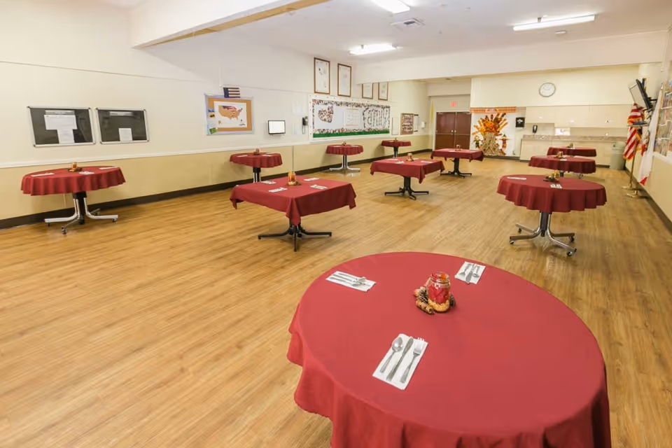 A spacious dining room with multiple round tables covered in red tablecloths, each set with utensils and a centerpiece. The room has wooden flooring, beige walls with bulletin boards and decorations, and an American flag in the corner.