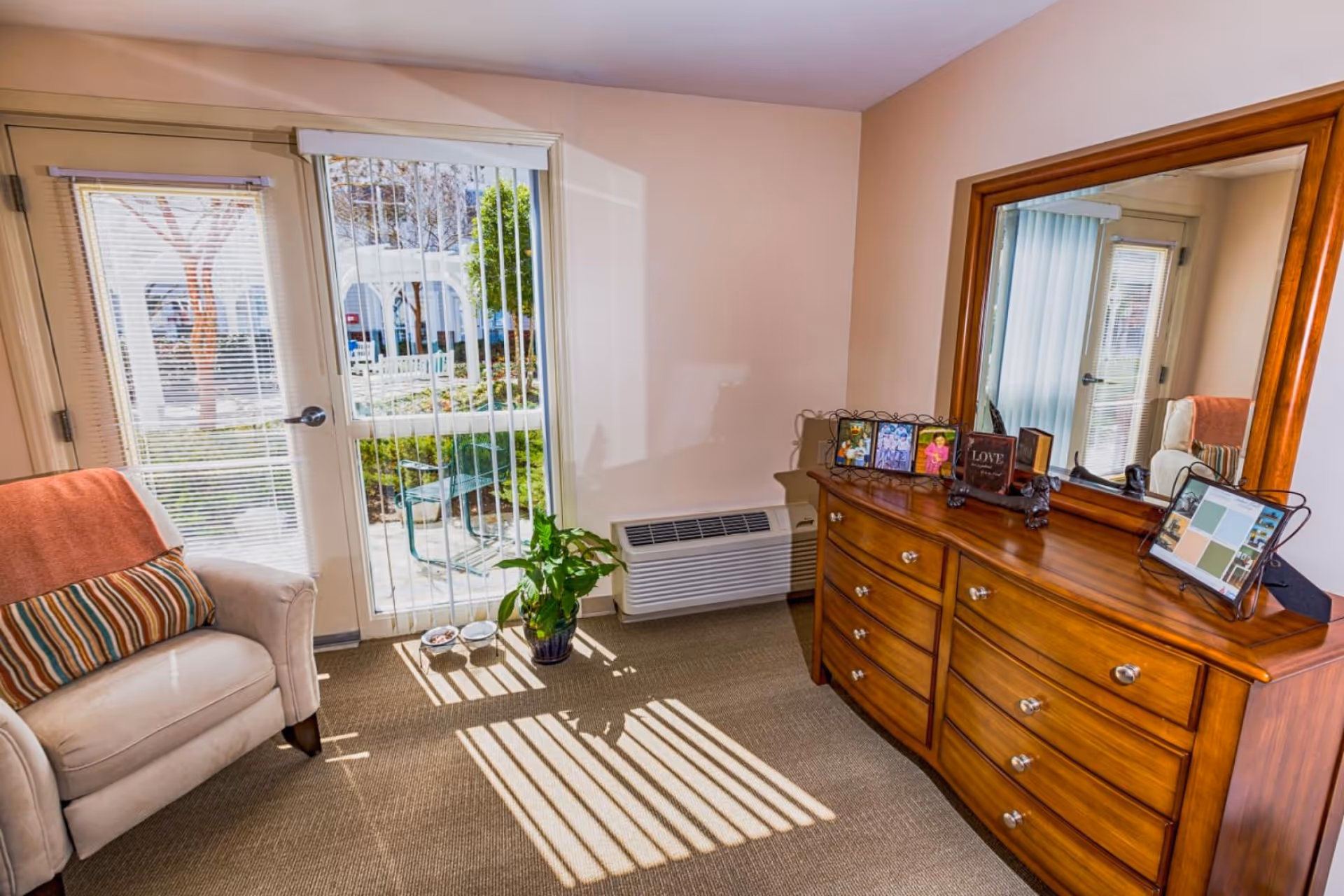 Sunlit bedroom corner with a wooden dresser and mirror, armchair, potted plant and sliding glass door to the outside.