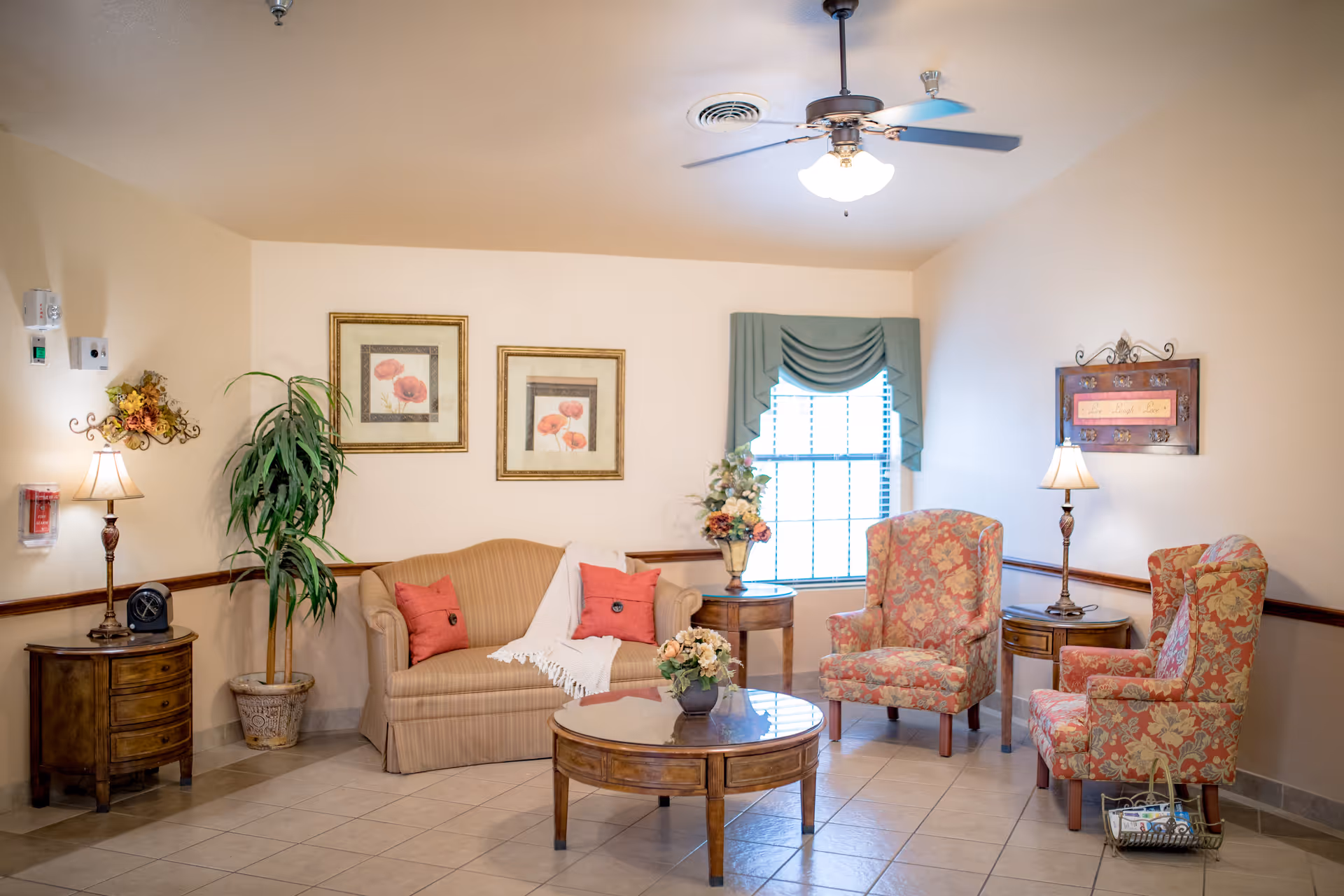 Cozy, well-lit sitting room with a sofa, two patterned armchairs, a round coffee table, lamps, framed artwork, and a window with drapery.