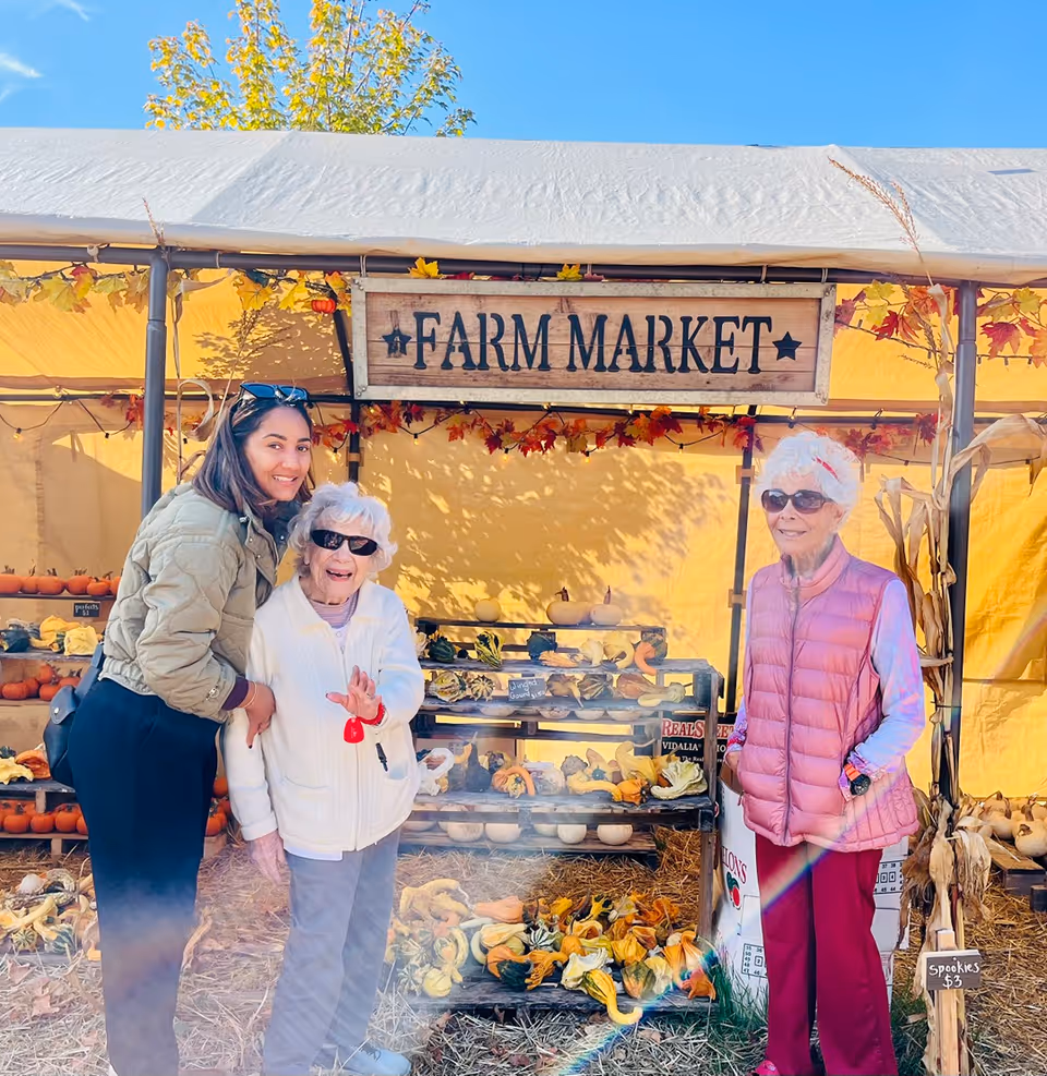 Three women smiling in front of a farm market stall displaying pumpkins and gourds under a canopy.
