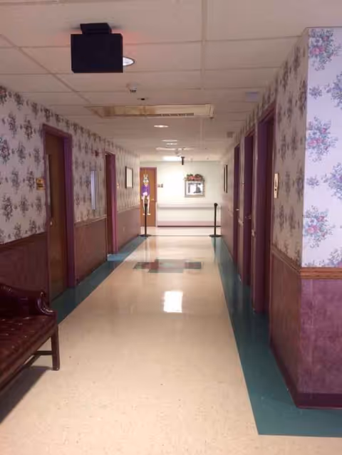 A long, brightly lit hallway inside a nursing home with floral wallpaper and a bench.