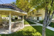 Sunlit landscaped courtyard featuring a white gazebo, paved walkways, green lawn, trees and a two-story residential building.