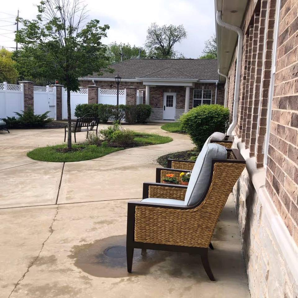 Outdoor courtyard with wicker patio chairs against a brick building, paved walkways, landscaped planting islands, a bench, and a small building in the background.