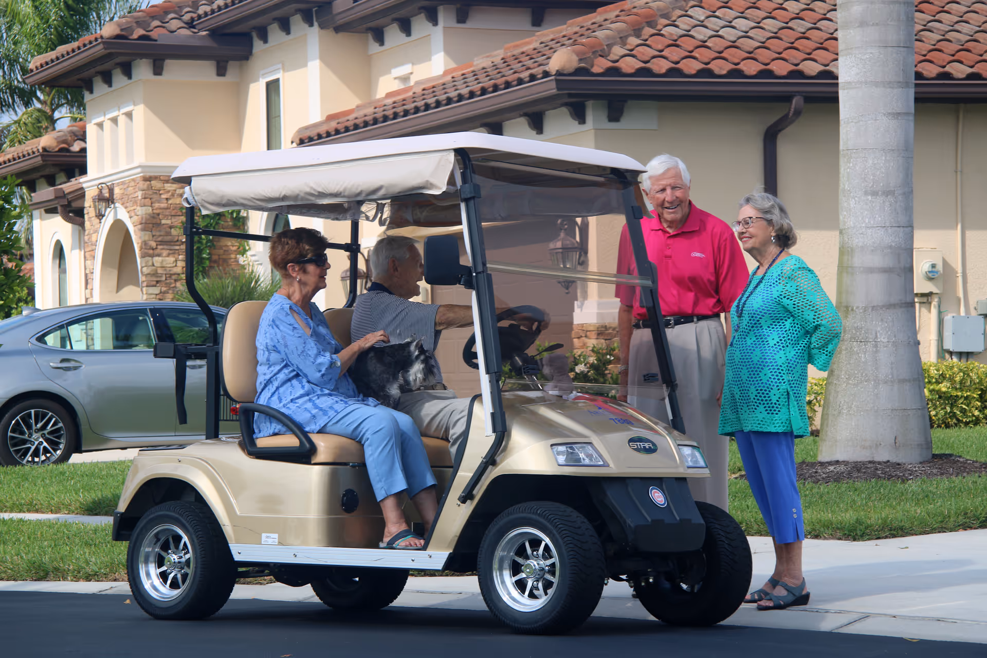 Two elderly women and one elderly man standing and talking next to a golf cart with another elderly man and woman seated inside it, in front of a residential building with a tiled roof and palm trees.
