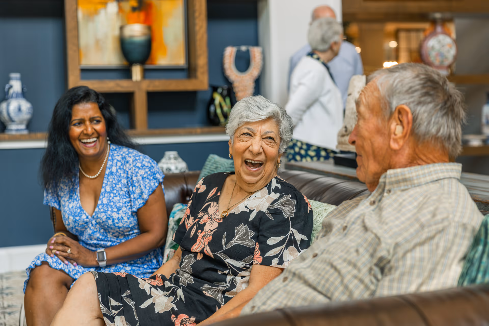 Three elderly people sitting on a brown leather couch in a living room, smiling and engaging in conversation. The background shows decorative items on shelves and two other people standing and talking.