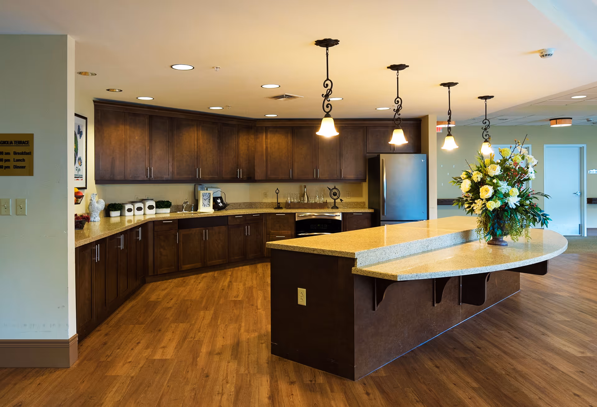 A spacious kitchen area with dark wooden cabinets, a large curved countertop island with a flower arrangement, pendant lights hanging from the ceiling, and a stainless steel refrigerator. The floor is wooden, and there is a sign on the wall indicating meal times.