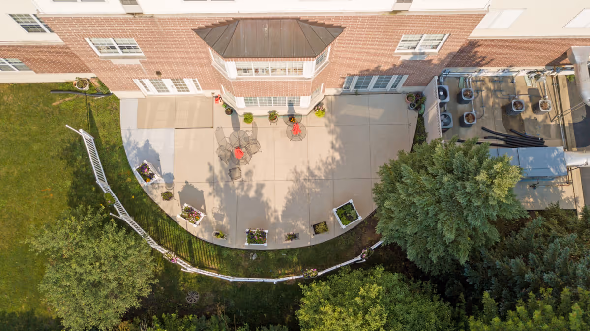 Aerial view of a patio area outside a brick building with several glass doors and windows. The patio has metal tables and chairs with red umbrellas, surrounded by potted plants and flower boxes. There is a white fence enclosing part of the grassy area with trees and shrubs nearby. HVAC units and piping are visible on the right side of the building.