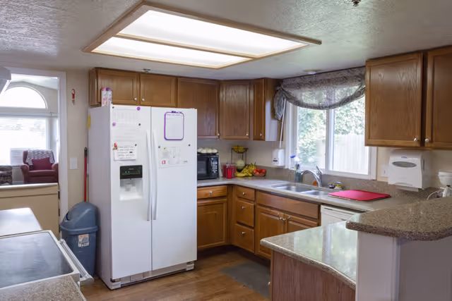 Bright kitchen with a white refrigerator, wooden cabinets, a sink under a window, and a speckled countertop/bar.