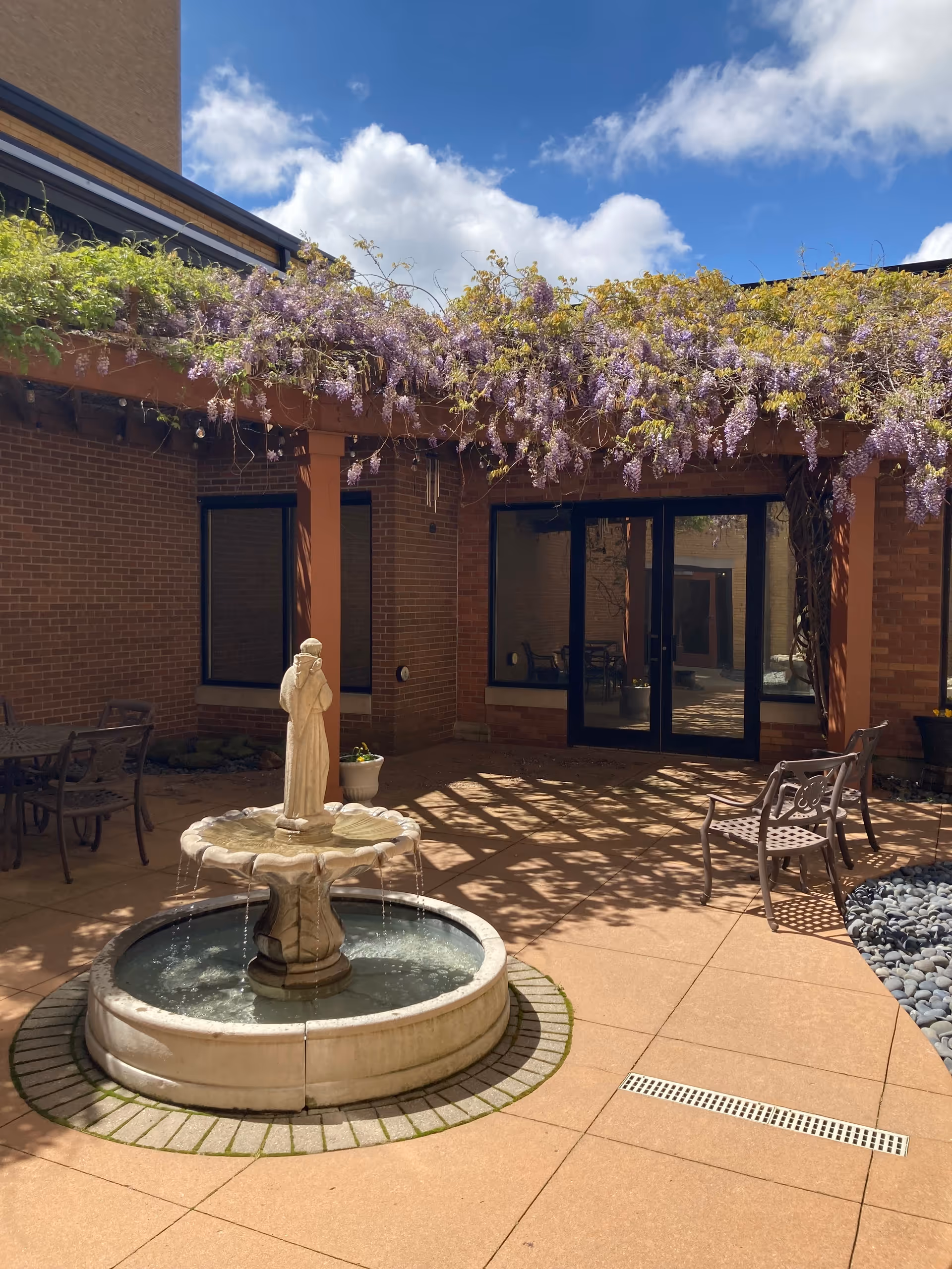 Outdoor courtyard area with a stone fountain featuring a statue in the center. The courtyard is surrounded by brick walls and has a pergola overhead covered with purple flowering vines. There are metal chairs and tables arranged around the courtyard, and the sky is blue with some clouds.