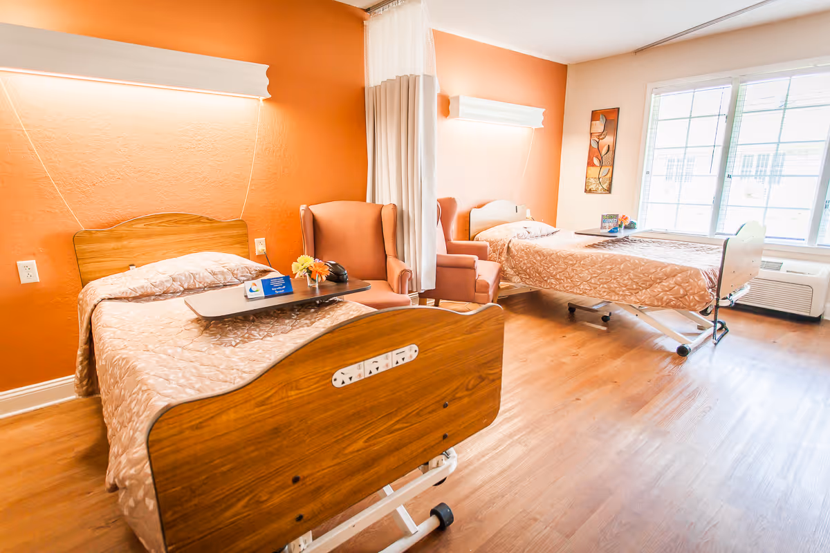 Two adjustable hospital beds in a bright senior living room with armchairs, a privacy curtain, and a large window.