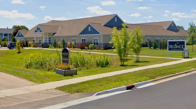 Exterior view of Willowick Assisted Living facility showing a single-story building with beige siding and a brown roof. The building is surrounded by green lawns, small trees, and shrubs. There are two signs visible, one near the sidewalk that says 'Willowick Entrance' and another larger sign further back that reads 'Willowick Assisted Living'. The sky is clear with a few clouds.
