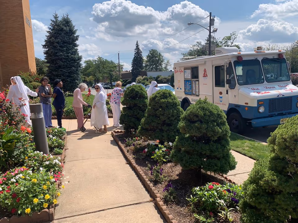 A group of elderly people and caregivers, including nuns, standing and walking on a sidewalk next to a garden with colorful flowers and trimmed bushes. A white ice cream truck is parked on the street nearby under a partly cloudy sky.