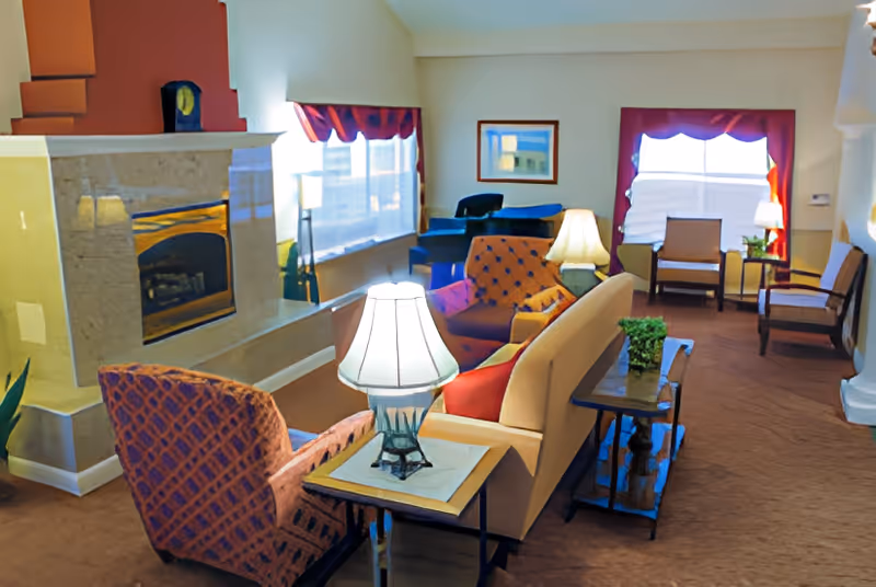 A cozy living room area with a beige sofa, patterned armchairs, a fireplace with a stone surround, two table lamps, a small side table with a plant, and windows with red curtains allowing natural light to enter.