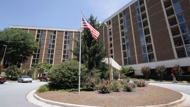Exterior view of a multi-story senior living facility with a circular driveway and landscaped center island featuring bushes, flowers, and an American flag on a flagpole.
