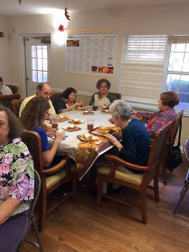 A group of elderly people sitting around a dining table in a well-lit room, eating a meal together. The table is covered with a white tablecloth and a decorative fall-themed runner. Plates of food and cups of drink are on the table. There is a window with blinds and a door in the background, along with a wall poster.