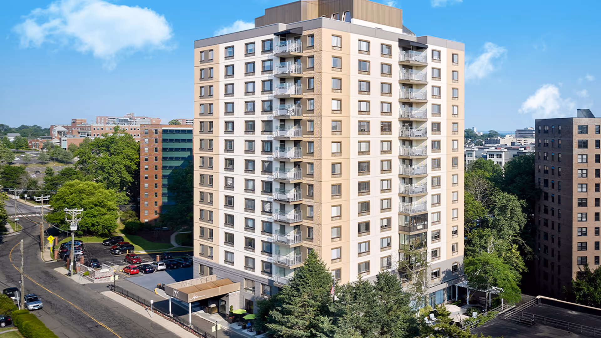 Tall beige apartment building with balconies in an urban setting surrounded by trees and nearby buildings under a blue sky.