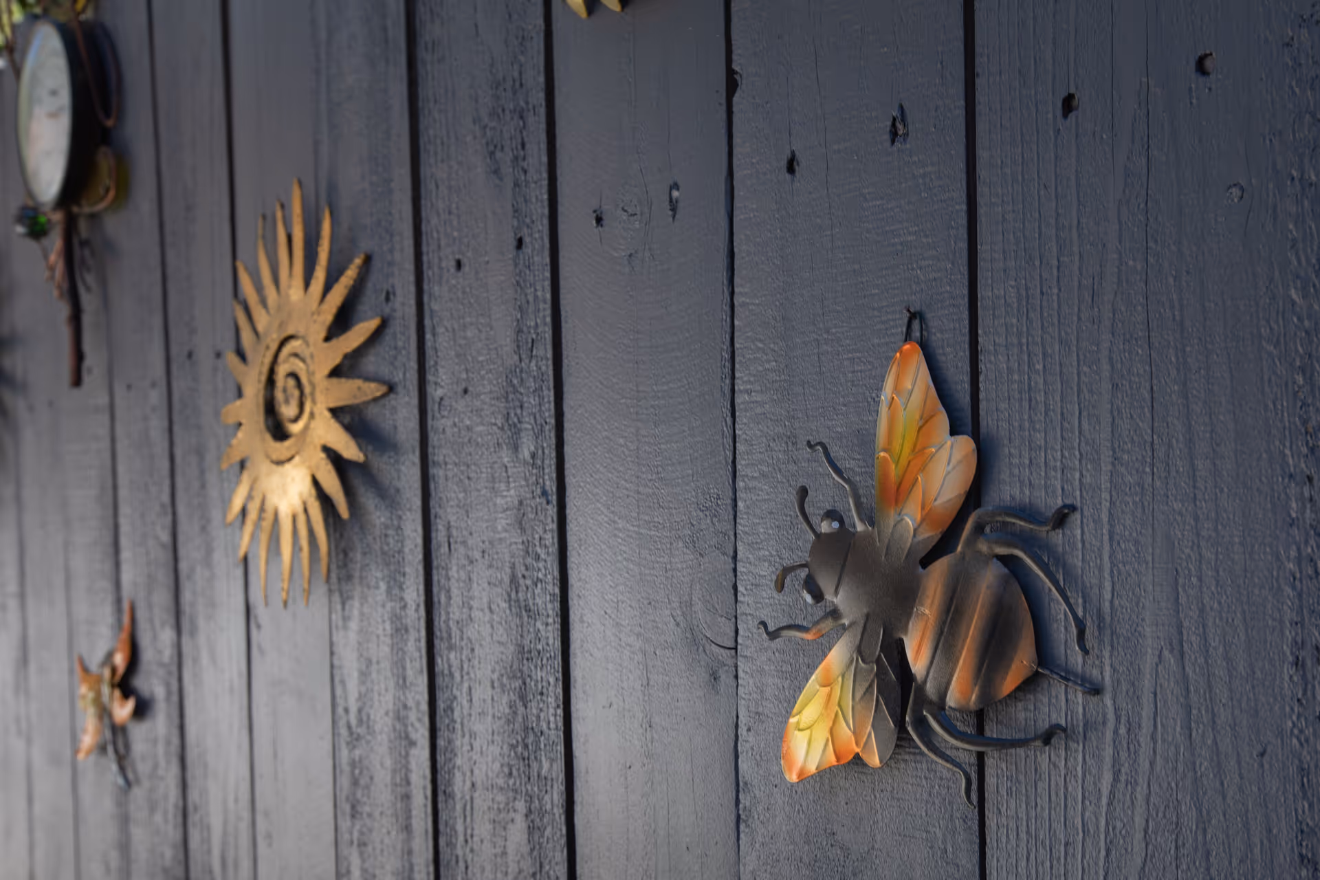 Decorative metal wall art including a large bee with orange and yellow wings, a golden sun, and other small metal ornaments mounted on a dark wooden wall.