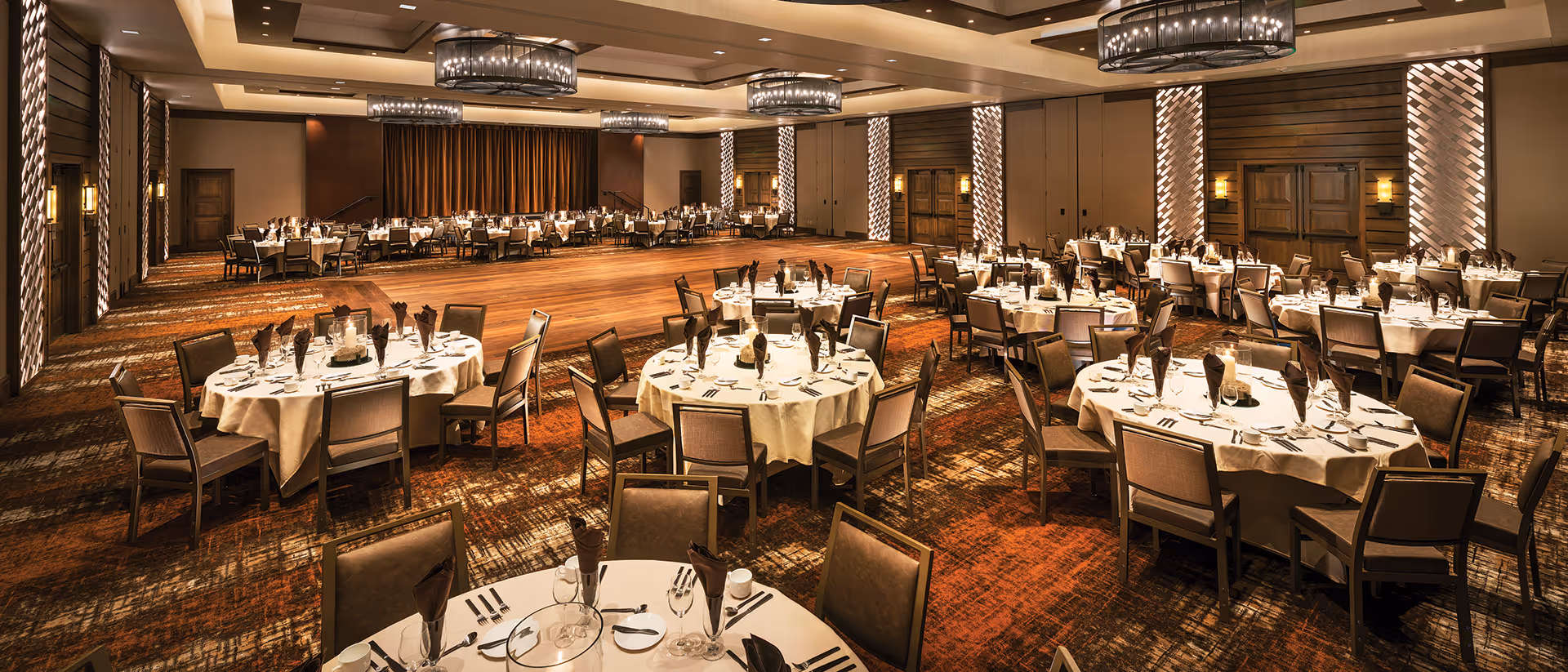 A large banquet-style dining hall with round tables set with linens, place settings, and chandeliers overhead.