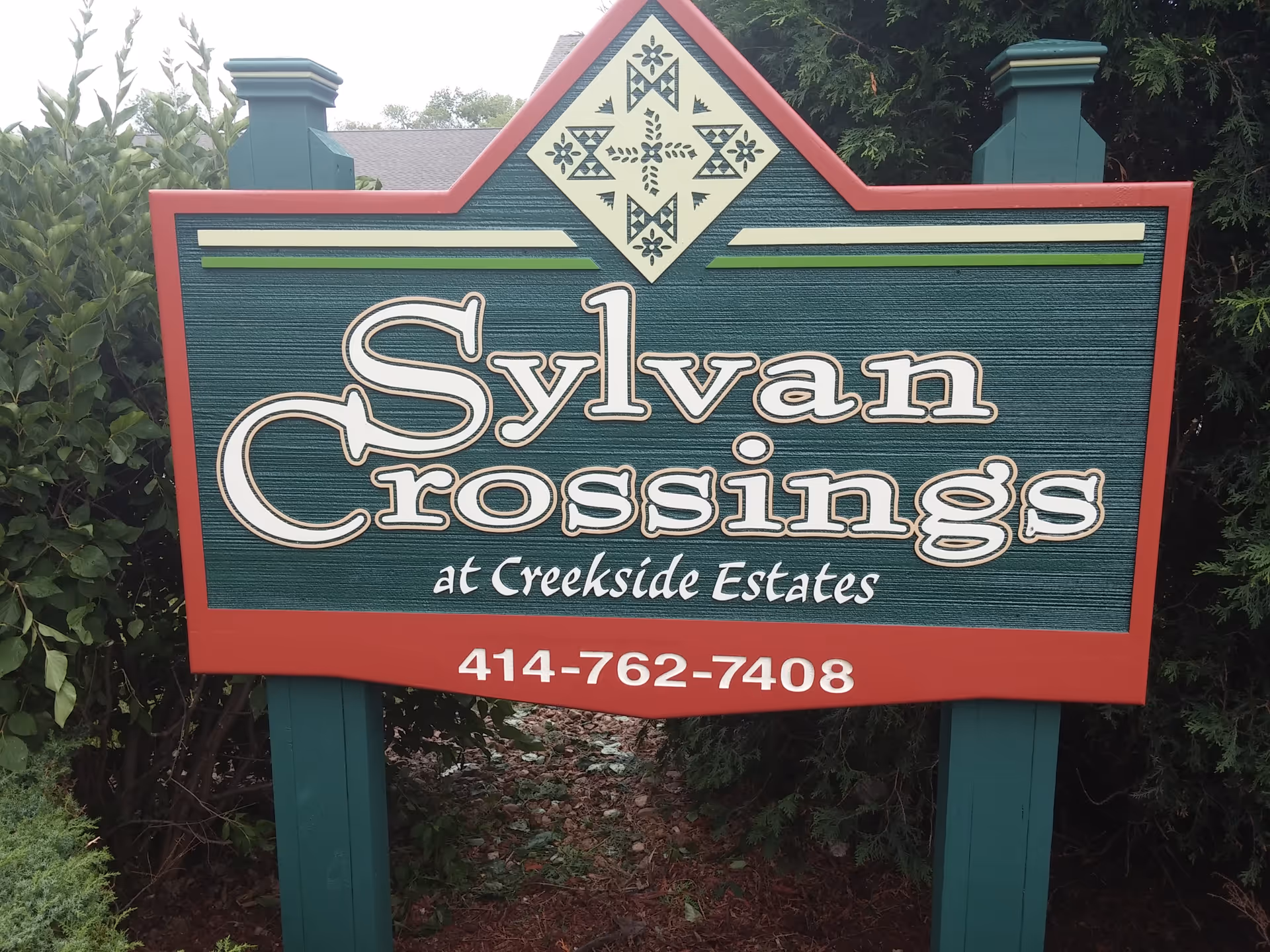 A decorative outdoor sign for Sylvan Crossings at Creekside Estates with a phone number 414-762-7408, surrounded by greenery.