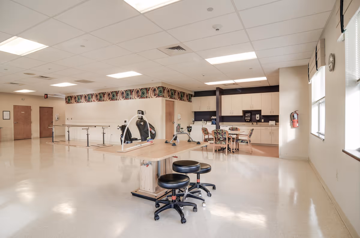 Spacious rehabilitation room with exercise equipment, parallel bars, a therapy table with stools, and a kitchenette area.