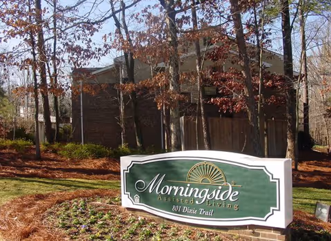 Outdoor view of the Morningside Assisted Living facility sign at 801 Dixie Trail, surrounded by trees and landscaping with a building partially visible in the background.