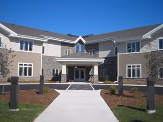 Front exterior view of a two-story senior living facility building with beige siding and stone accents, a covered entrance, multiple windows, and a clear blue sky above. There is a paved walkway leading to the entrance, flanked by landscaped areas with small shrubs and decorative posts.