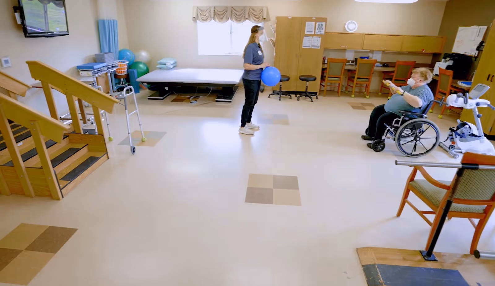 A rehabilitation room with a woman holding a blue exercise ball standing in front of a man in a wheelchair who is reaching out towards her. The room has exercise equipment including stairs with handrails, a walker, therapy balls, a treatment table, chairs, and a desk area with a computer. Both individuals are wearing masks.