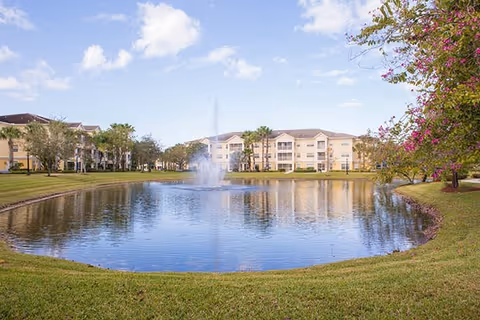 A landscaped pond with a central fountain in front of a multi-story senior living building under a partly cloudy sky.