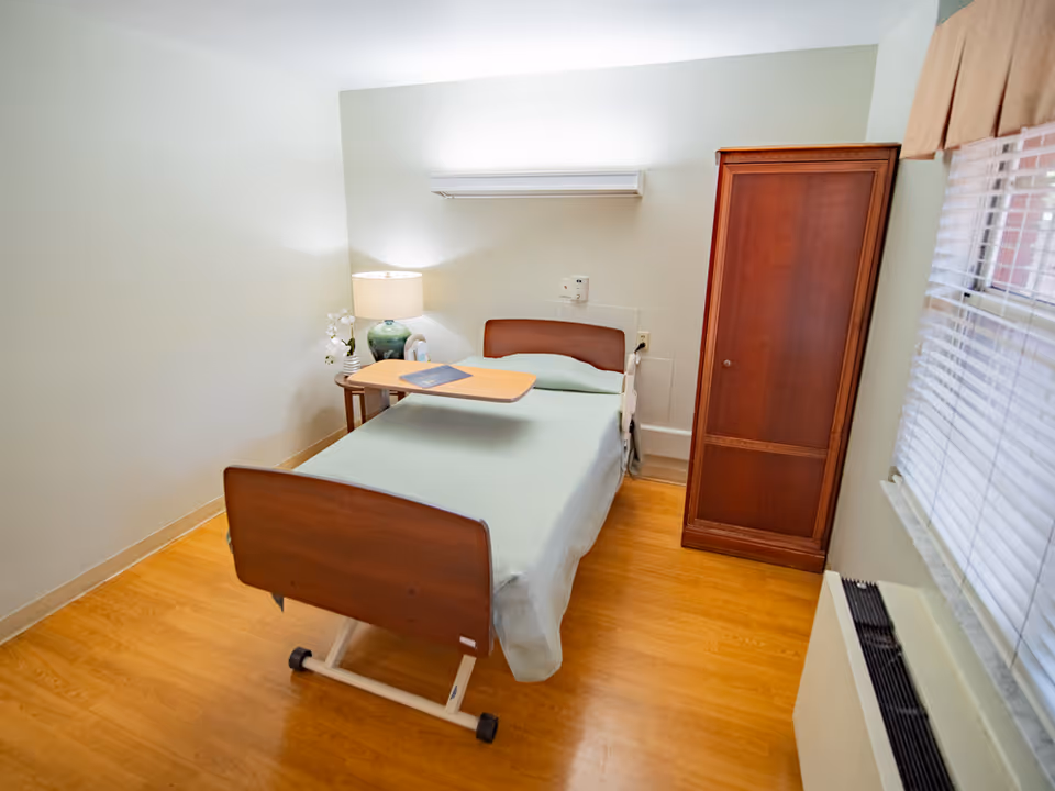 A small patient bedroom in a senior living facility with a single bed, overbed table, lamp, and a tall wooden wardrobe.