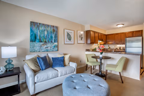 A cozy living area with a light gray sofa adorned with blue and beige cushions, a round tufted ottoman in front, and a small black side table with a blue glass lamp. Behind the sofa, there are three framed artworks on the wall. To the right, there is a small dining area with a round black table and two light green upholstered chairs. The background features a kitchen with wooden cabinets, a granite countertop, and stainless steel refrigerator.