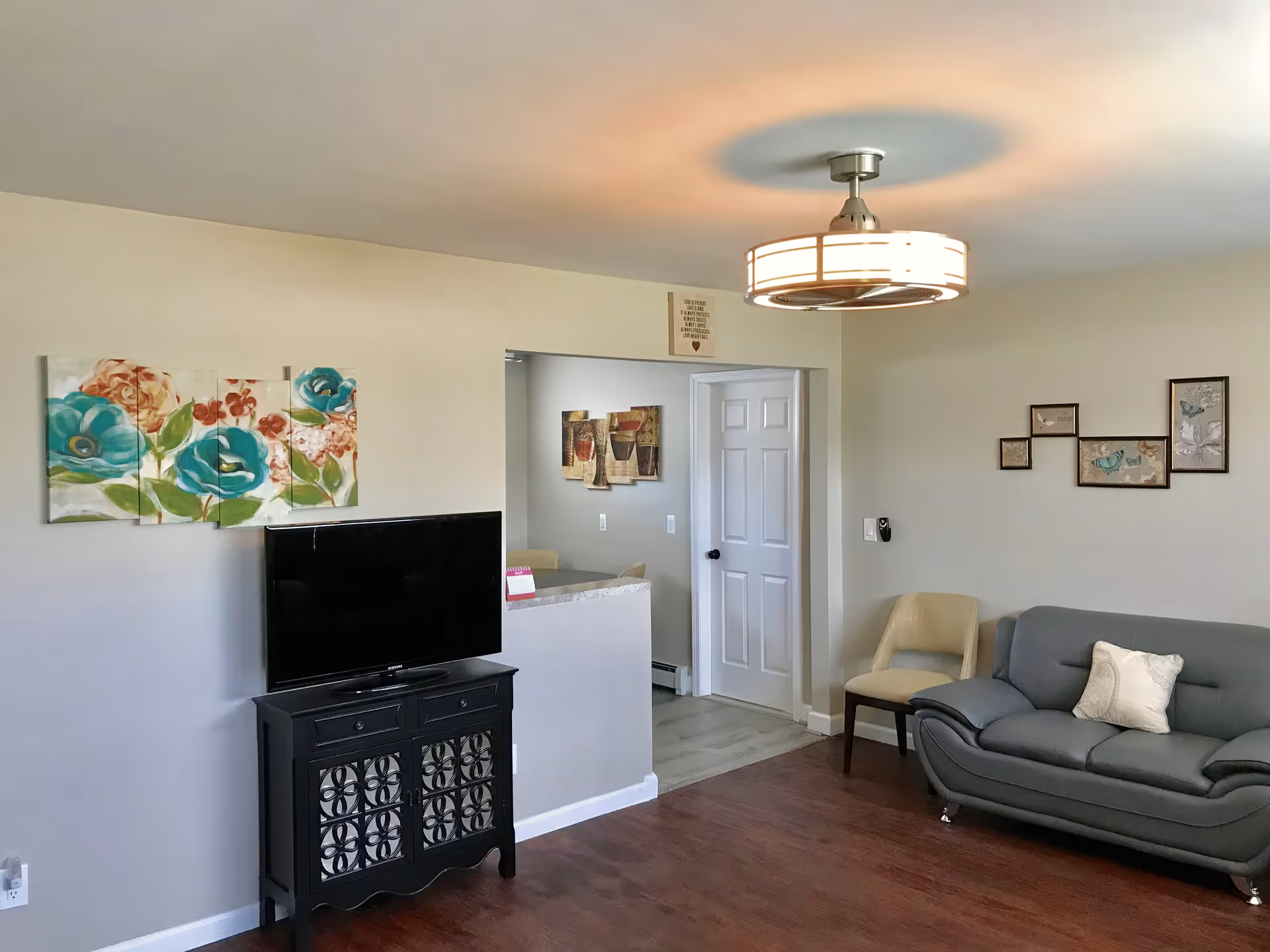 A cozy living room with a gray sofa, a beige chair, a black TV stand with a flat-screen TV, floral wall art, and a modern ceiling light fixture. The room has wooden flooring and light-colored walls. A doorway leads to another room with additional artwork visible.