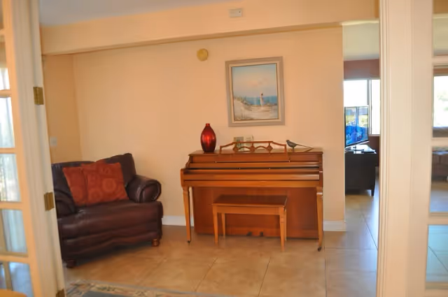Interior living area with an upright piano and bench, a leather armchair with red pillows, and a framed seaside painting on the wall.