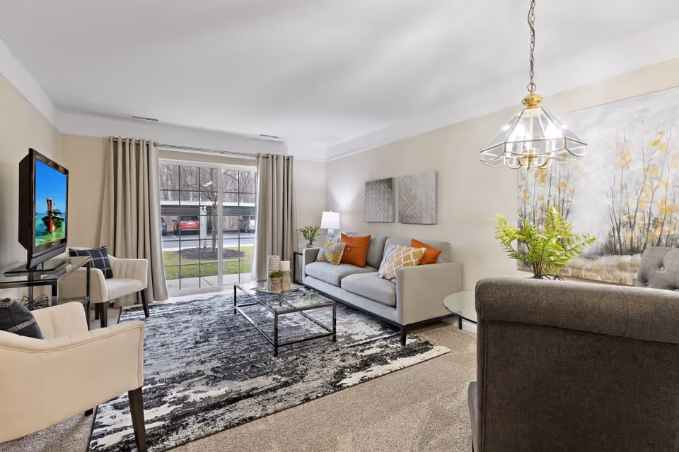 A bright and cozy living room with a large window and sliding glass door showing an outdoor parking area. The room features a gray sofa with orange and patterned cushions, two beige armchairs, a glass coffee table on a patterned rug, a side table with a lamp, and a large wall painting with floral designs. A glass chandelier hangs from the ceiling.