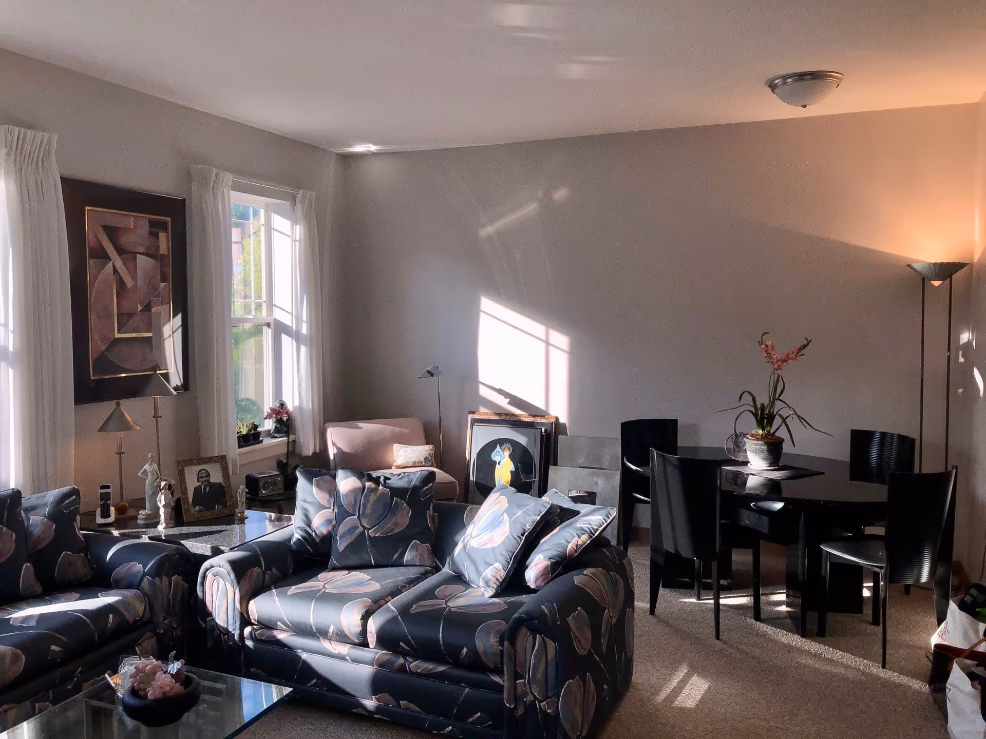 A cozy living room with floral patterned sofas, a glass coffee table, and a dining area with a round black table and four black chairs. The room is softly lit by natural light coming through two windows with white curtains. There are decorative items including framed pictures, a floor lamp, and a potted plant on the dining table.