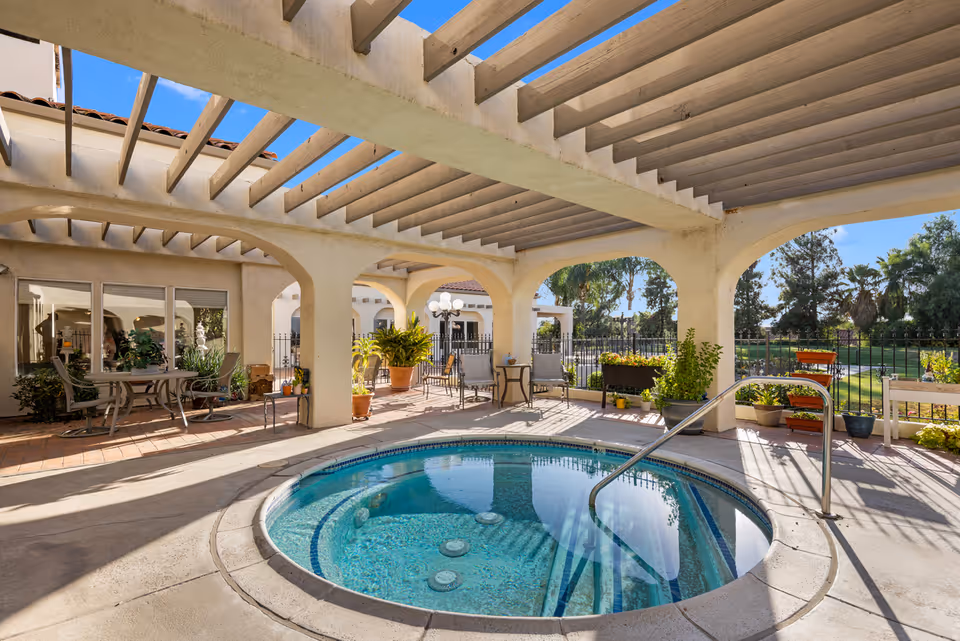 Outdoor covered patio area with a round hot tub in the center, surrounded by potted plants and patio furniture including tables and chairs. The area is shaded by a pergola-style roof with beams, and there is a view of a green lawn and trees beyond a black metal fence.