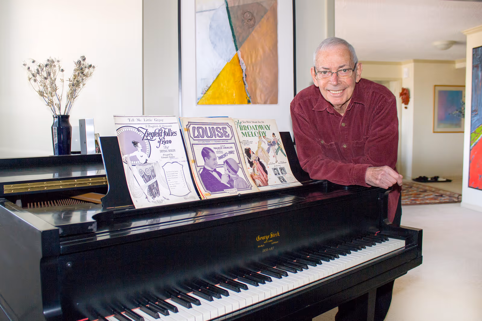 An elderly man wearing glasses and a red shirt leans on a black grand piano with sheet music displayed on the music stand. The room has light-colored walls, modern artwork, and a vase with dried flowers on the piano.
