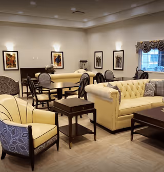 A well-lit living room area with beige tufted sofas, patterned armchairs, dark wooden tables, framed artwork on the walls, and a window with a decorative valance.
