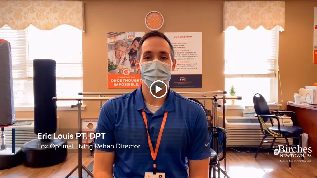A man wearing a blue polo shirt and a face mask sits in a rehabilitation room with exercise equipment and chairs. Behind him are two windows with blinds, a clock, and a poster on the wall. Text on the image identifies him as Eric Louis PT, DPT, Fox Optimal Living Rehab Director. The Birches at Newtown logo is visible in the bottom right corner.