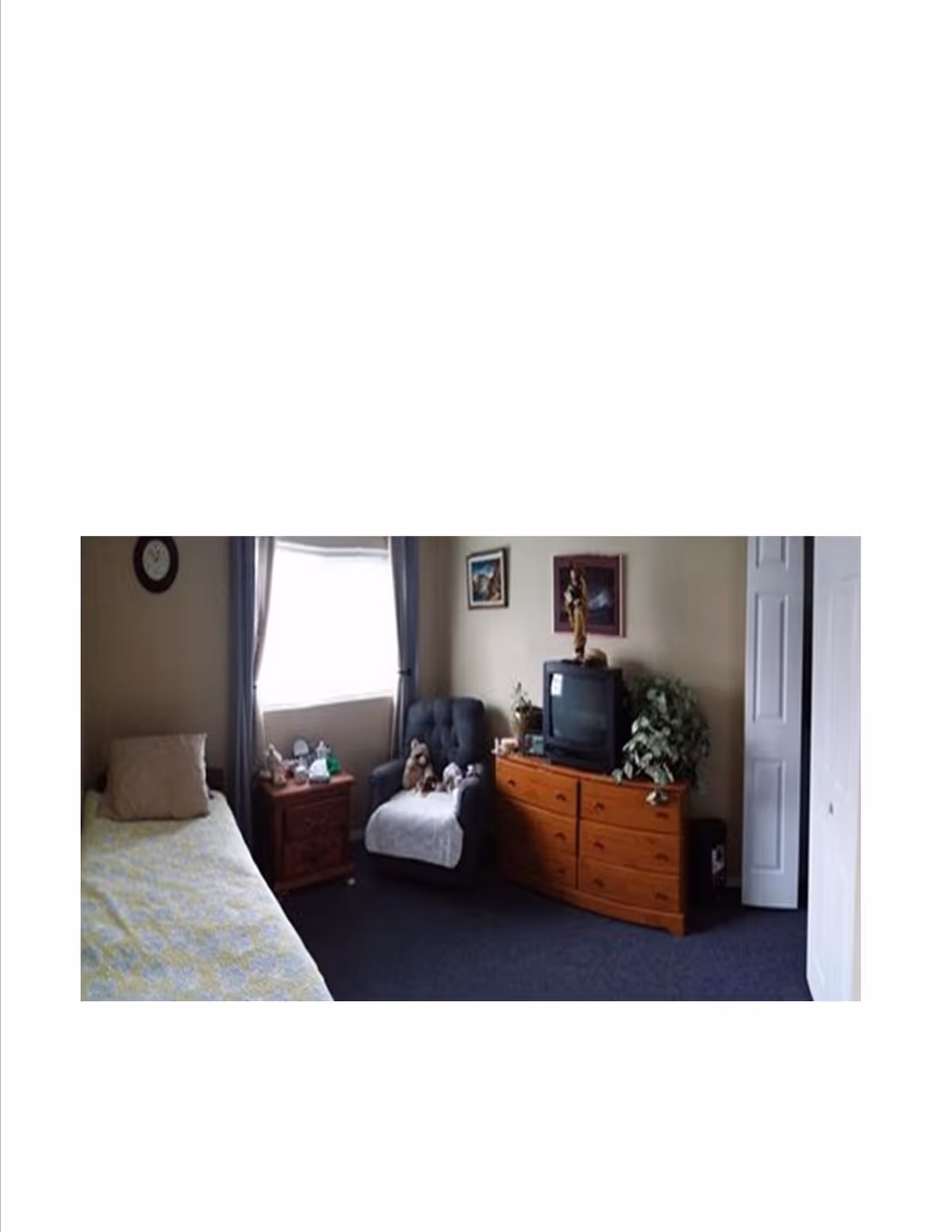 A cozy bedroom with a single bed covered in a light-colored quilt, a wooden nightstand with various items, a cushioned armchair with stuffed animals, a wooden dresser with a TV and decorative items, a plant, and framed pictures on the walls. A window with curtains lets in natural light.