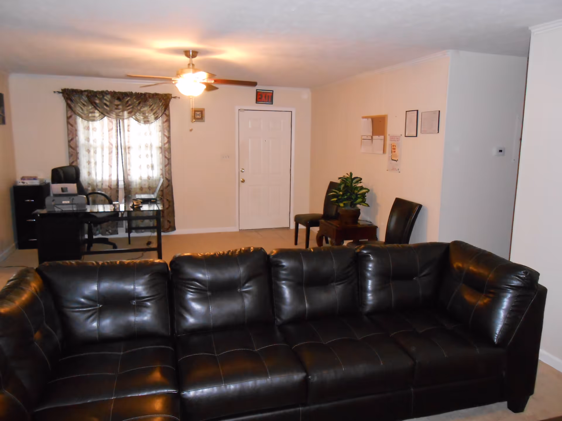 Interior view of a living room with a black leather sectional sofa in the foreground. In the background, there is a desk with a chair, printer, and laptop near a window with patterned curtains. Two chairs and a small table with a potted plant are placed against a wall with some papers and frames. A ceiling fan with a light is on the ceiling, and a white door with an exit sign above it is visible.