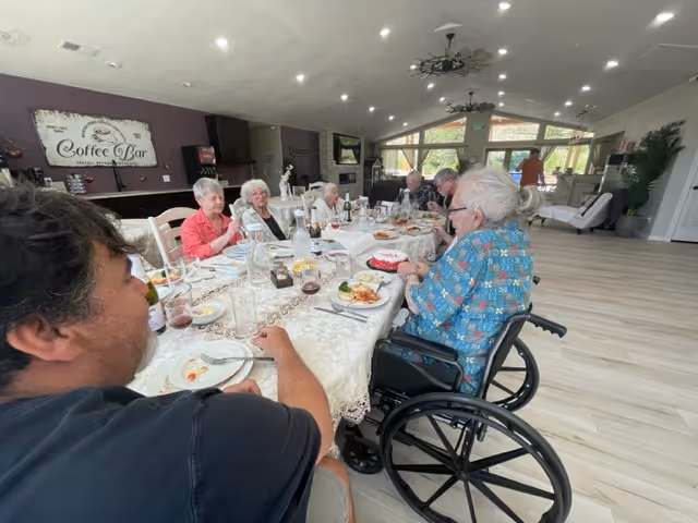 A group of elderly people and a younger man sitting around a long dining table in a spacious, well-lit room. The table is set with plates of food, glasses, and a cake. One elderly woman is in a wheelchair. The room has large windows, light wooden floors, and a sign on the wall that reads 'Coffee Bar'.