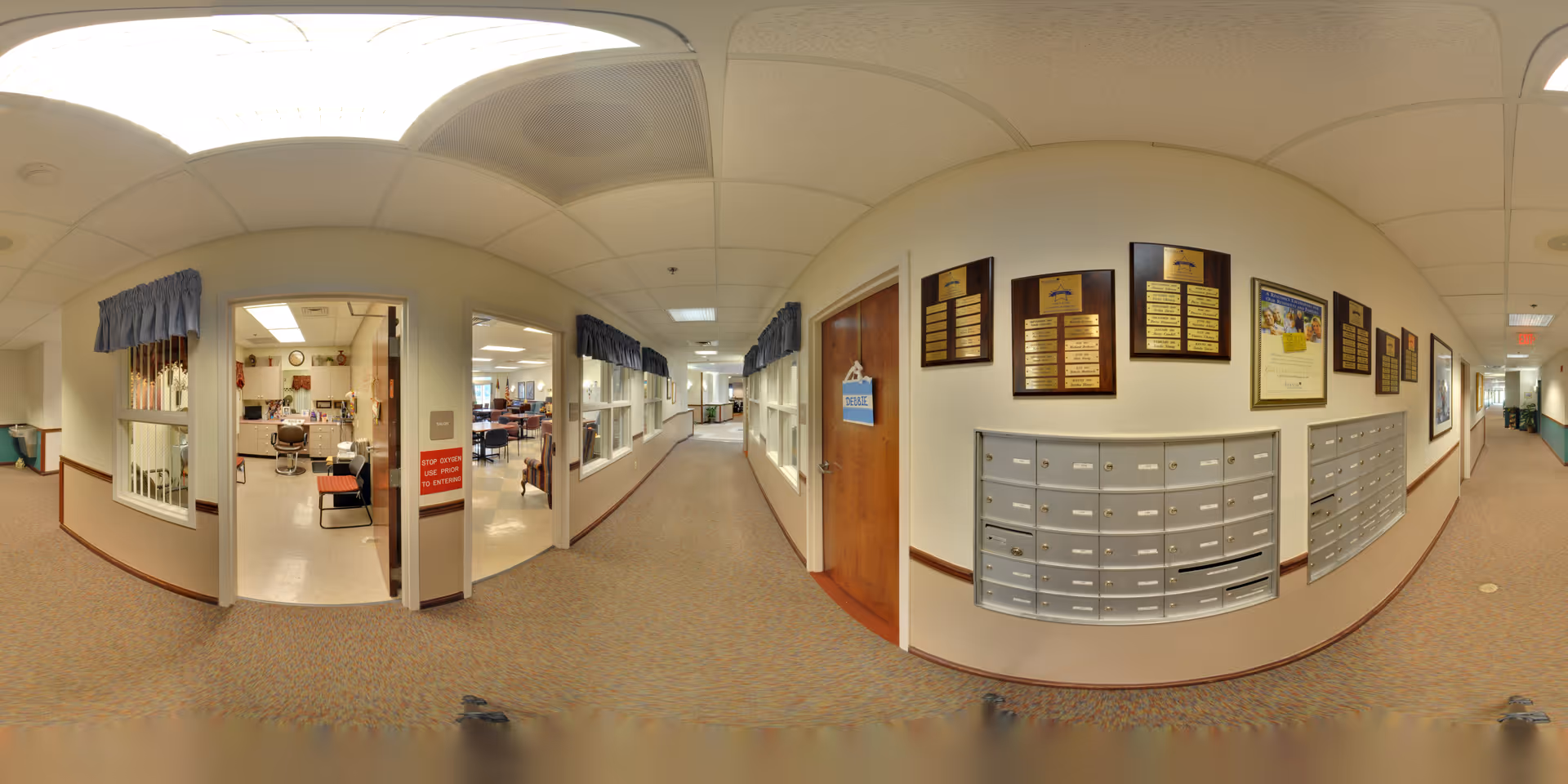 Carpeted interior hallway of a senior living facility showing wall-mounted mailboxes and plaques, windows into activity rooms, and a long corridor.
