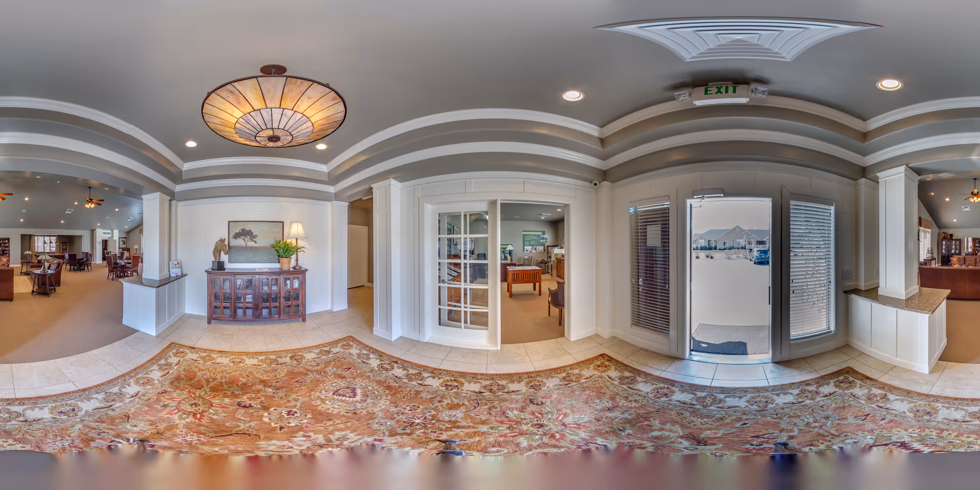 Wide panoramic view of the interior entrance area of Bridlewood Villas senior living facility, featuring a decorative ceiling light, a patterned area rug, a wooden cabinet with a lamp and plant, glass doors leading to an office or meeting room, and a glass exit door with windows on either side showing an outdoor parking area.