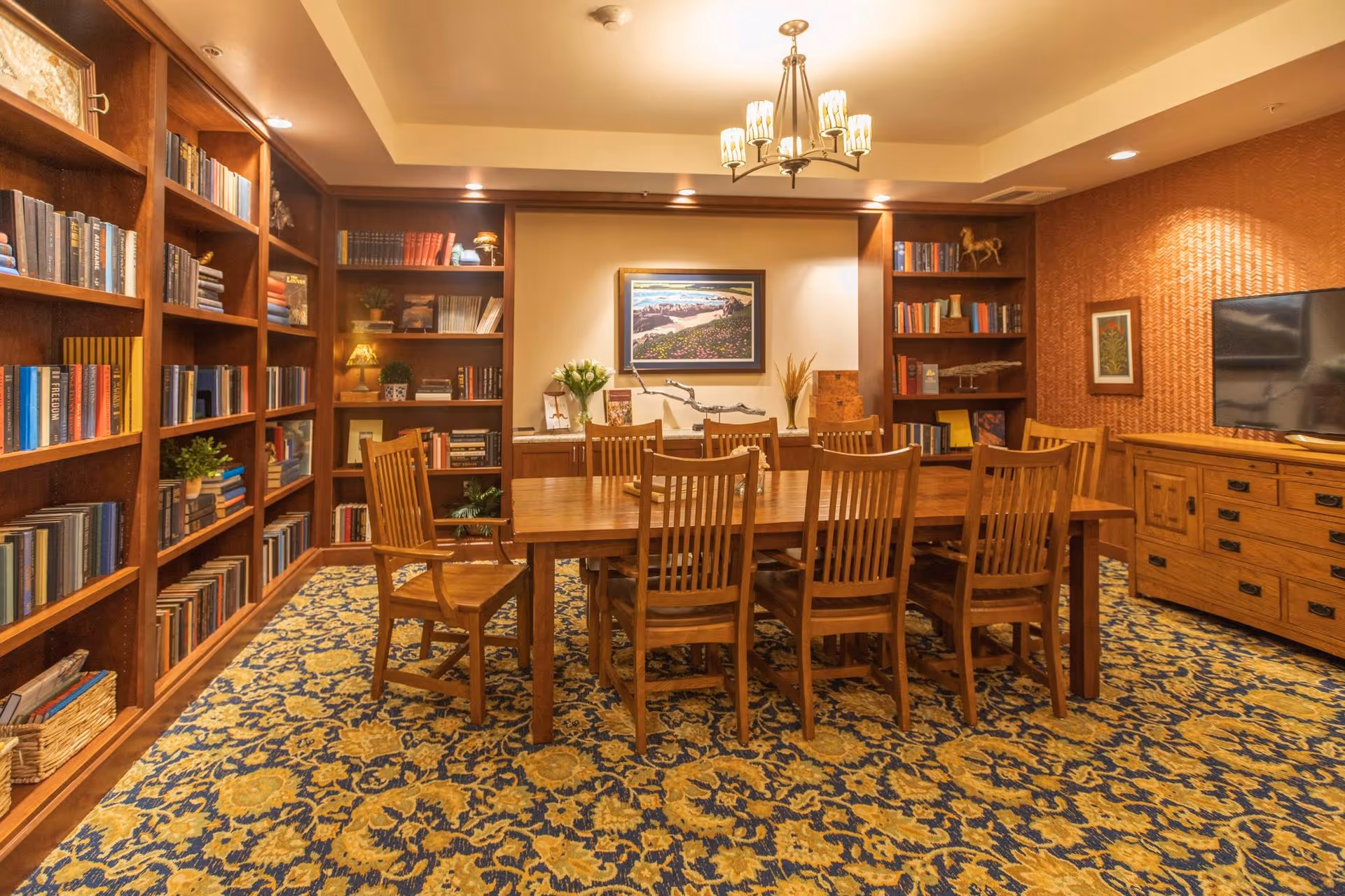 A cozy library or reading room with wooden bookshelves filled with books lining the walls. A large wooden table with eight matching chairs is centered on a patterned carpet. The room is warmly lit by a chandelier and recessed lighting, with framed artwork and decorative items on the shelves and walls.